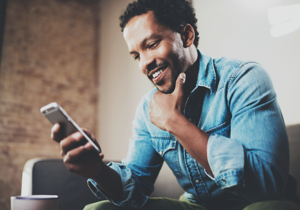 Man in a blue denim shirt smiling and rubbing his chin while reading on his smartphone.