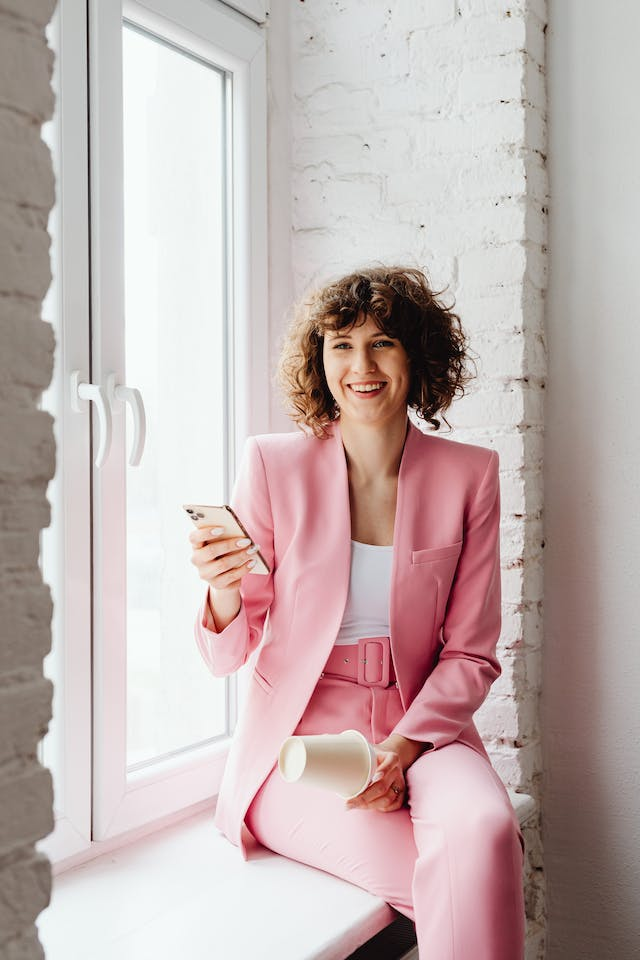Woman with pink business outfit sitting on windowsill holding phone and smiling