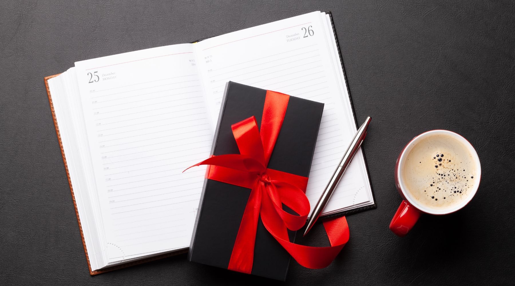 Black gift box with red ribbon on an open planner with a cup of coffee, indicating a scheduled gift-giving