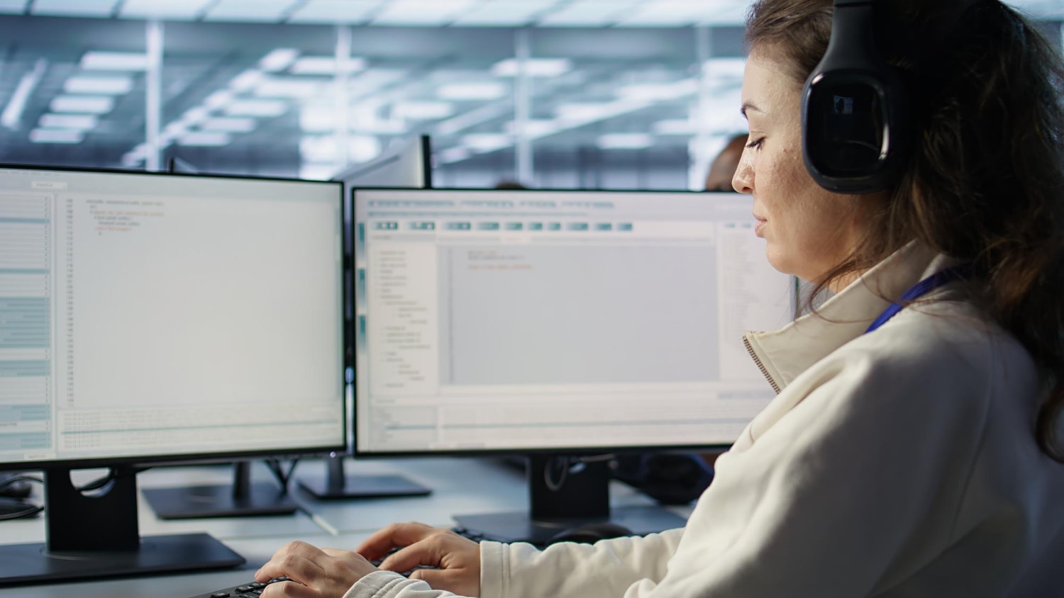 A woman with a headset types on her keyboard while monitoring business phone system data on multiple screens. The scene illustrates VoIP, call routing, and customer communication tools.