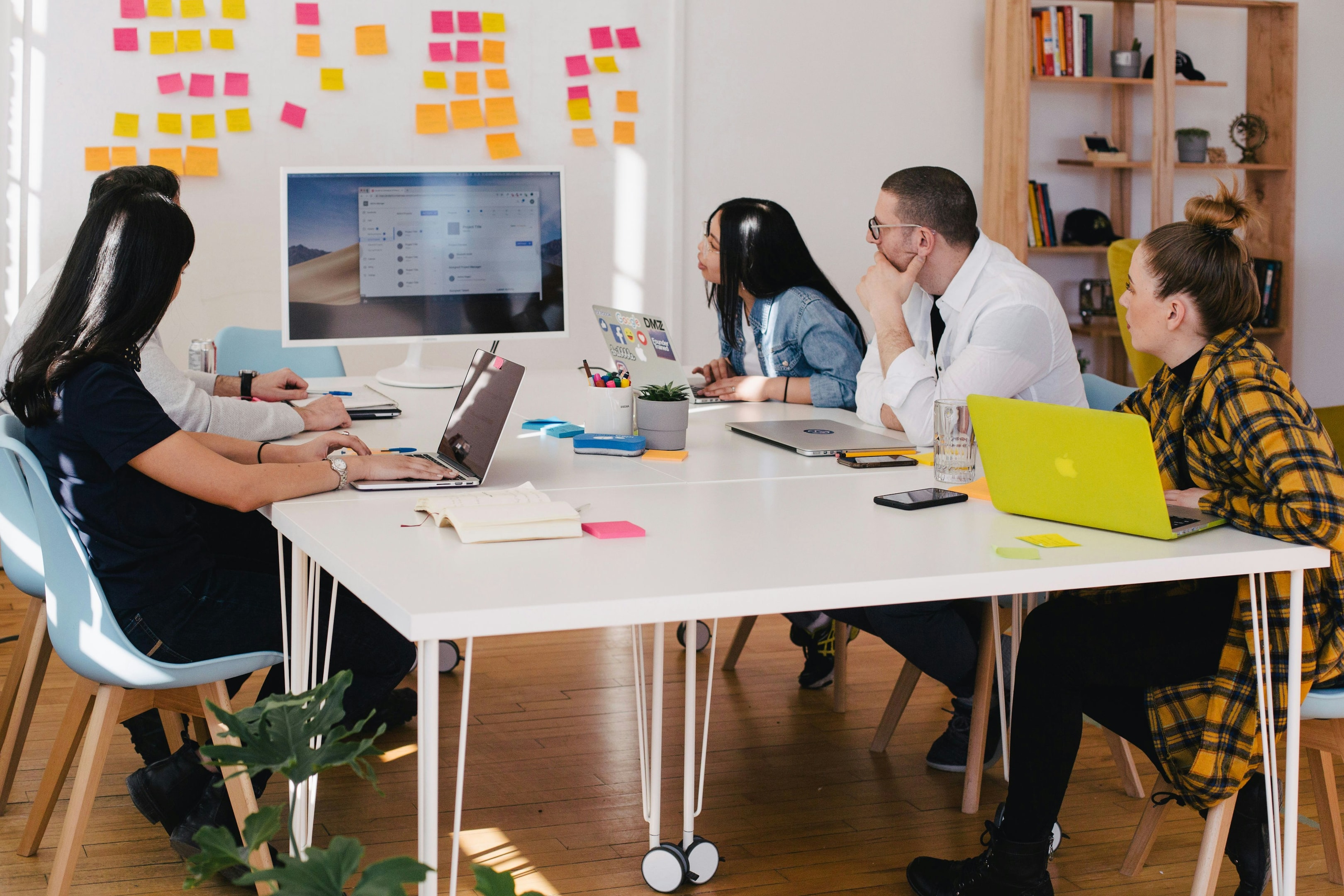 Five people at a table looking at a screen in front of a board with sticky notes.