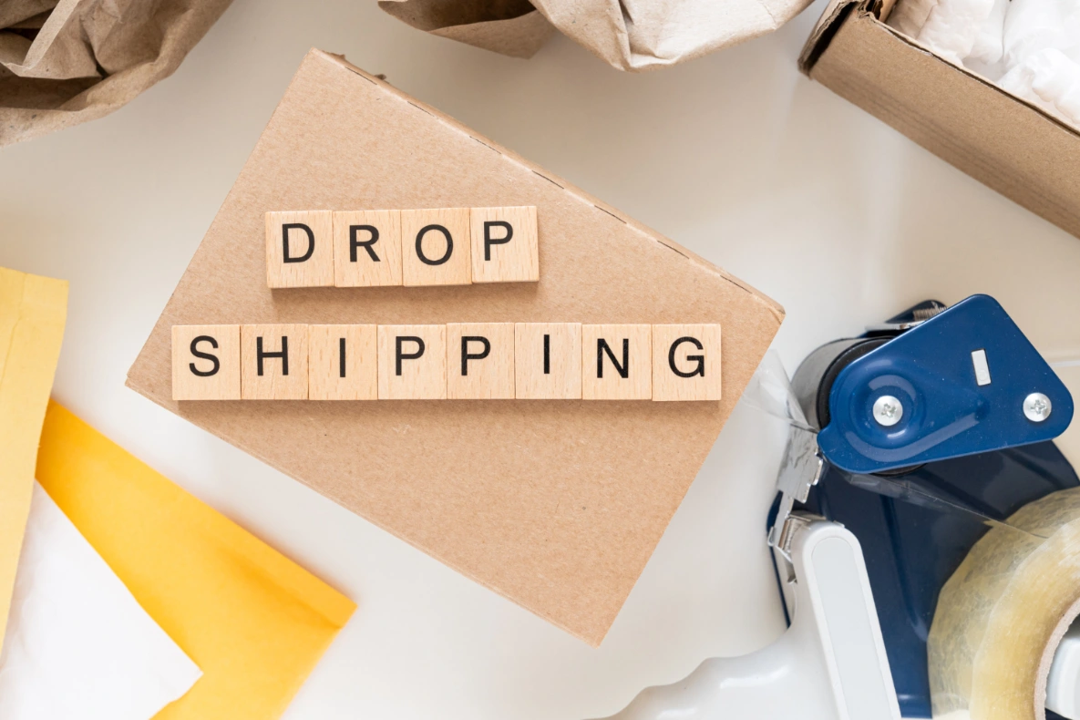 Wooden blocks spelling out "DROP SHIPPING" rest on a cardboard box surrounded by packing materials and a tape dispenser.