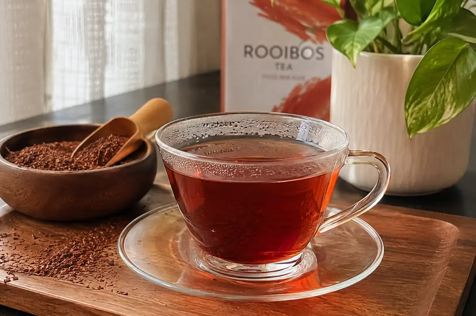 Arranged on a dark wooden tray, a clear glass teacup filled with deep reddish-orange brewed rooibos tea sits on its matching saucer. Next to the cup, a small wooden bowl holds dry rooibos tea leaves with a tiny wooden scoop resting inside, surrounded by a light dusting of loose leaves on the tray, with a potted houseplant and a tea box softly blurred in the background.