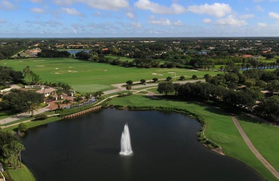 The image depicts a championship golf course in Quail West, Naples, featuring pristine fairways and scenic water hazards, all basking under the bright Florida sunshine. This tranquil golf community offers an exceptional lifestyle with immediate golf membership access and beautifully landscaped surroundings.