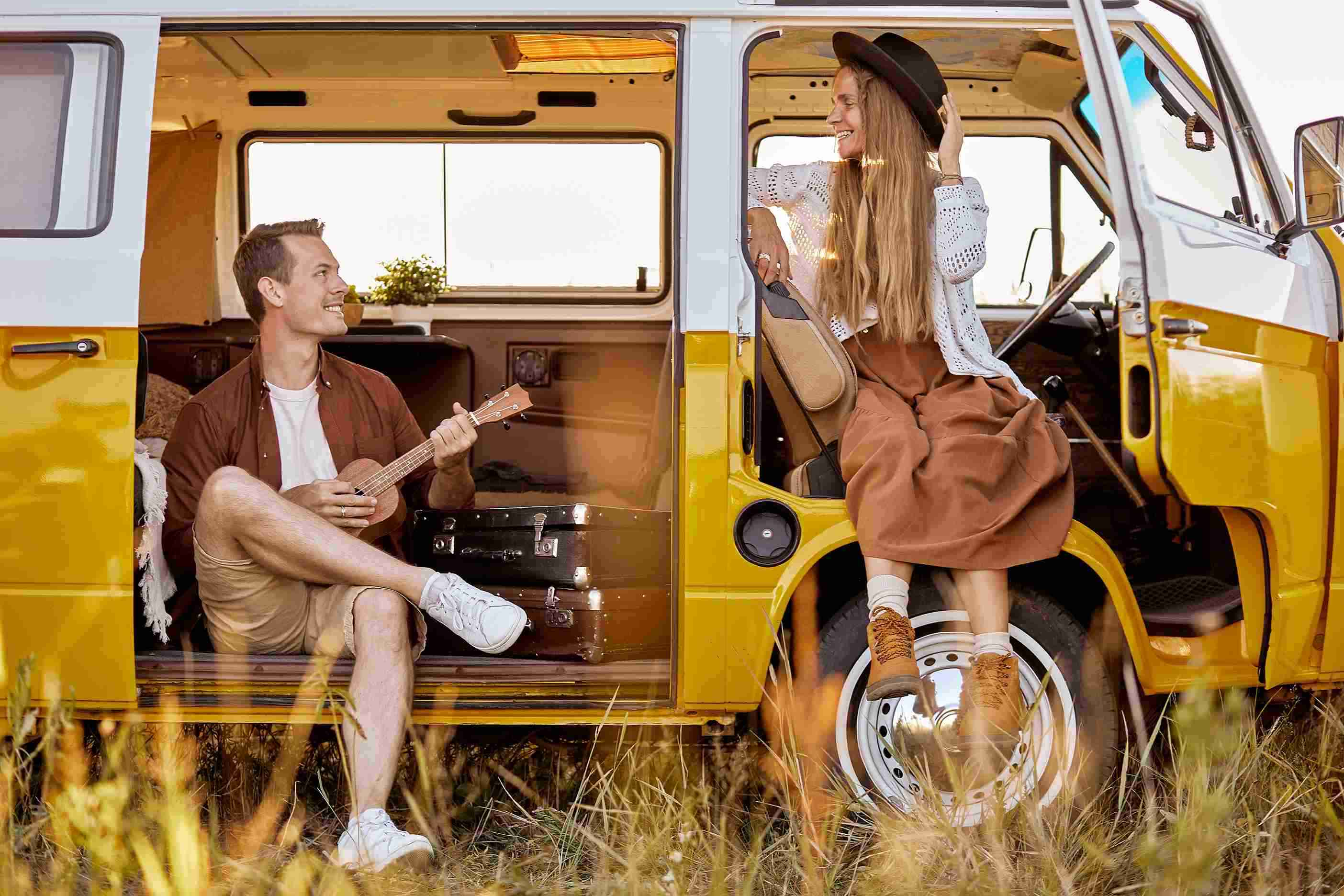 Casal hipster feliz, homem a tocar ukulele, a cantar uma canção numa campervan amarela no campo