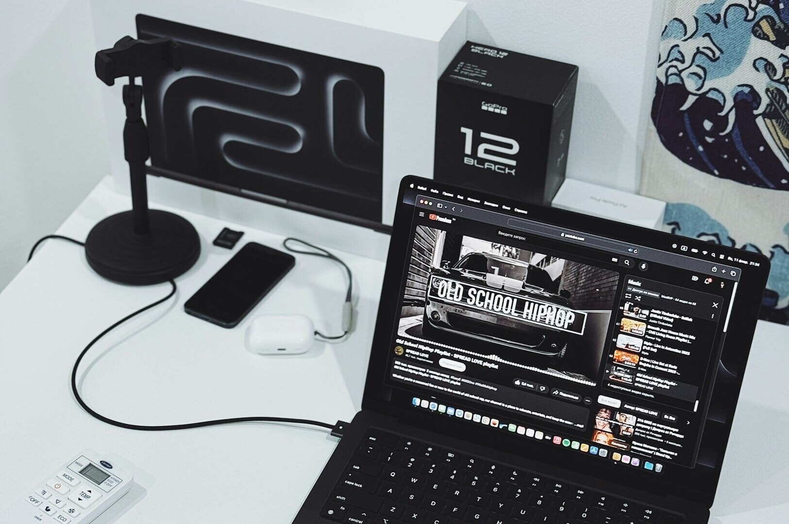 A modern workspace with a black laptop playing "Old School Hip Hop." Nearby, a smartphone, earbuds, and a microphone are on a white desk, conveying a tech-savvy vibe.