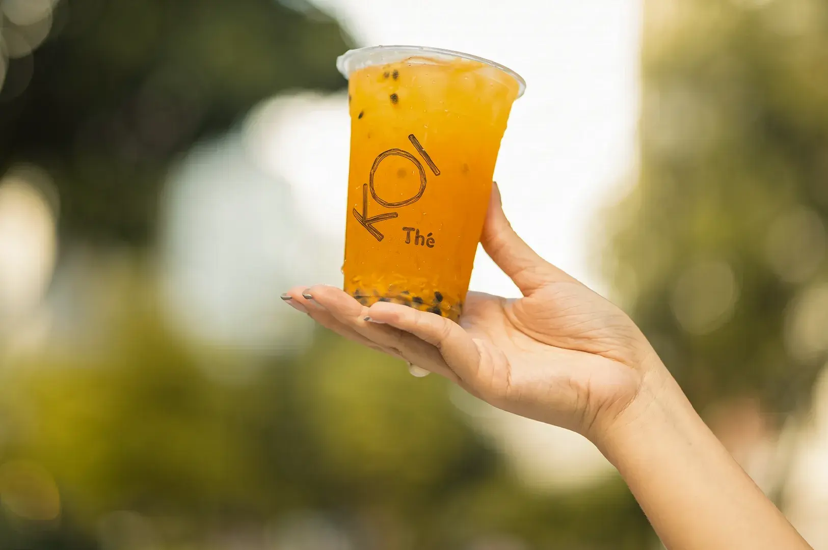 Hand holding a branded KOI Thé iced passion fruit tea cup against a soft-focus outdoor background.
