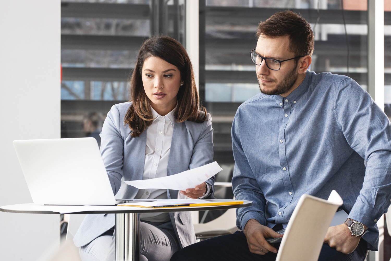 Two people at a table, one working on a laptop and the other holding papers.