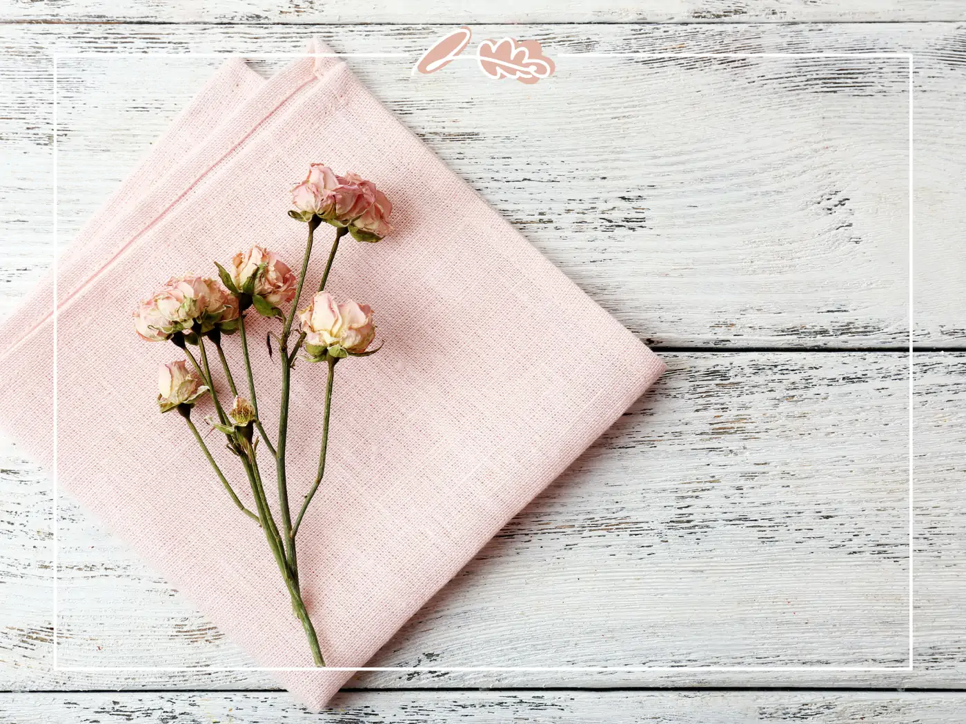 A sprig of dried pink roses placed on a folded pink napkin, set against a rustic white wooden table. Fabulous Flowers and Gifts.