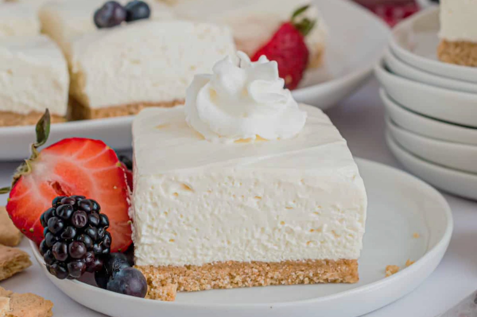 A creamy cheesecake slice with graham cracker crust topped with whipped cream, garnished with sliced strawberry, blackberry, and blueberries on a white plate.