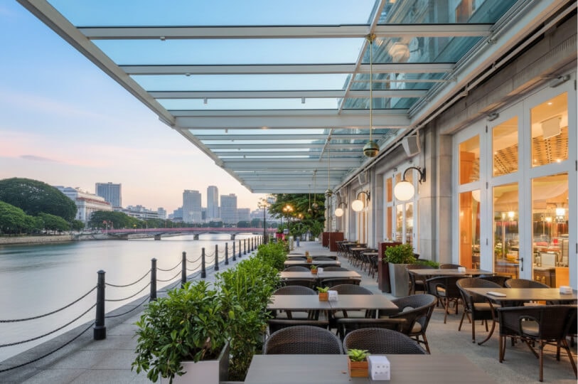 A scenic riverside patio at dusk with a modern glass awning covering several outdoor dining tables. The walkway is lined with green shrubs and a black railing, overlooking a calm river and a city skyline with a bridge in the distance.