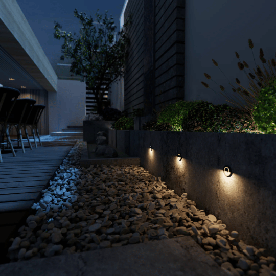 Modern outdoor pathway with small recessed wall lights illuminating stone landscaping and plants at night beside a seating area.