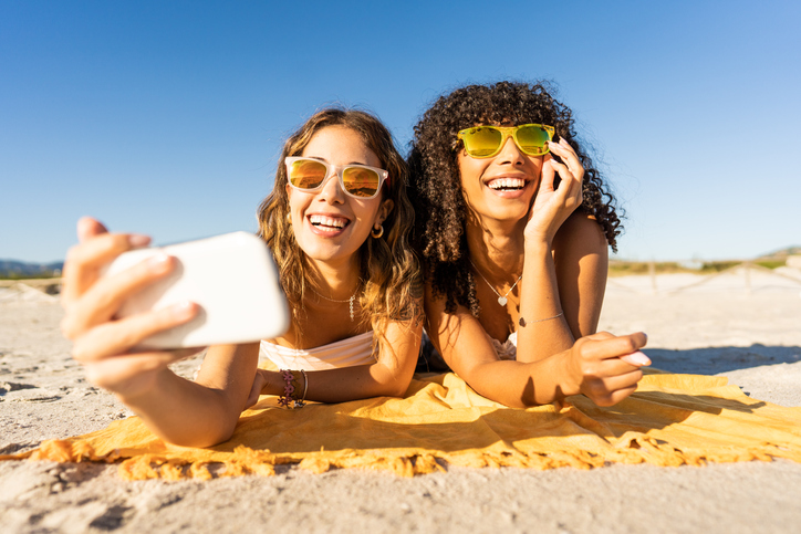 Two happy young women laying on the beach wearing sunglasses and looking at a cellphone.  