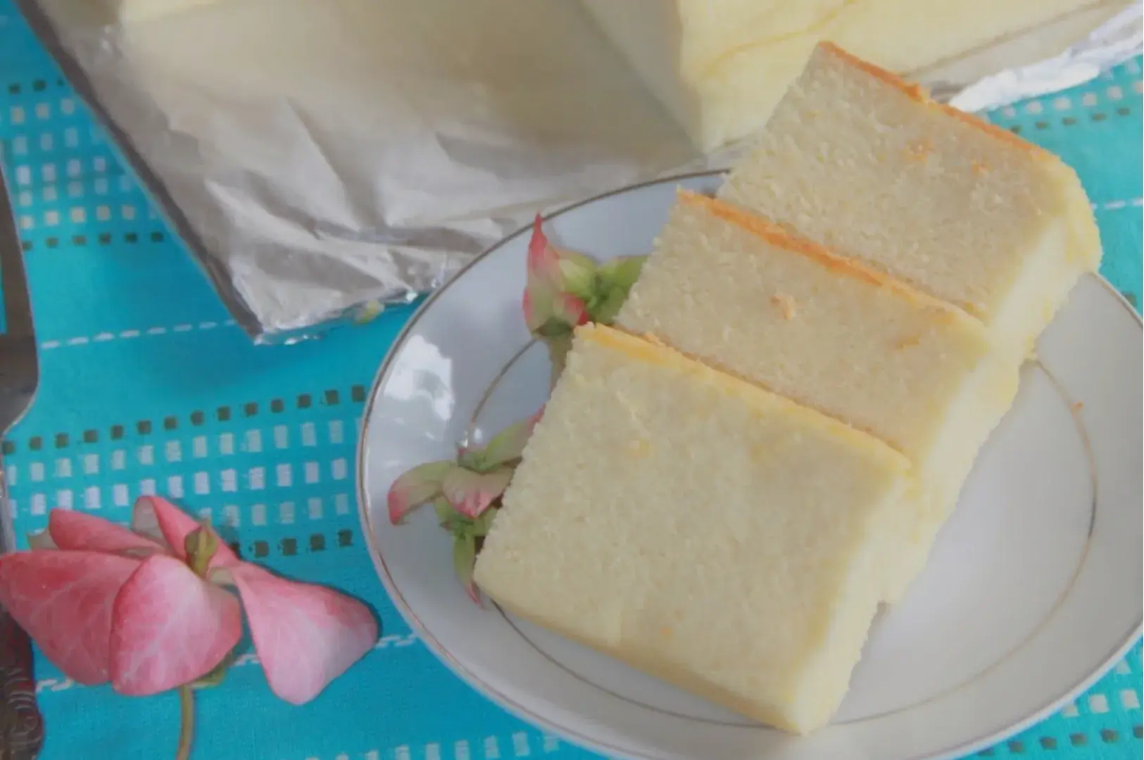 A slice of cake on a plate with flowers beside it, with a larger piece of cake in the background.