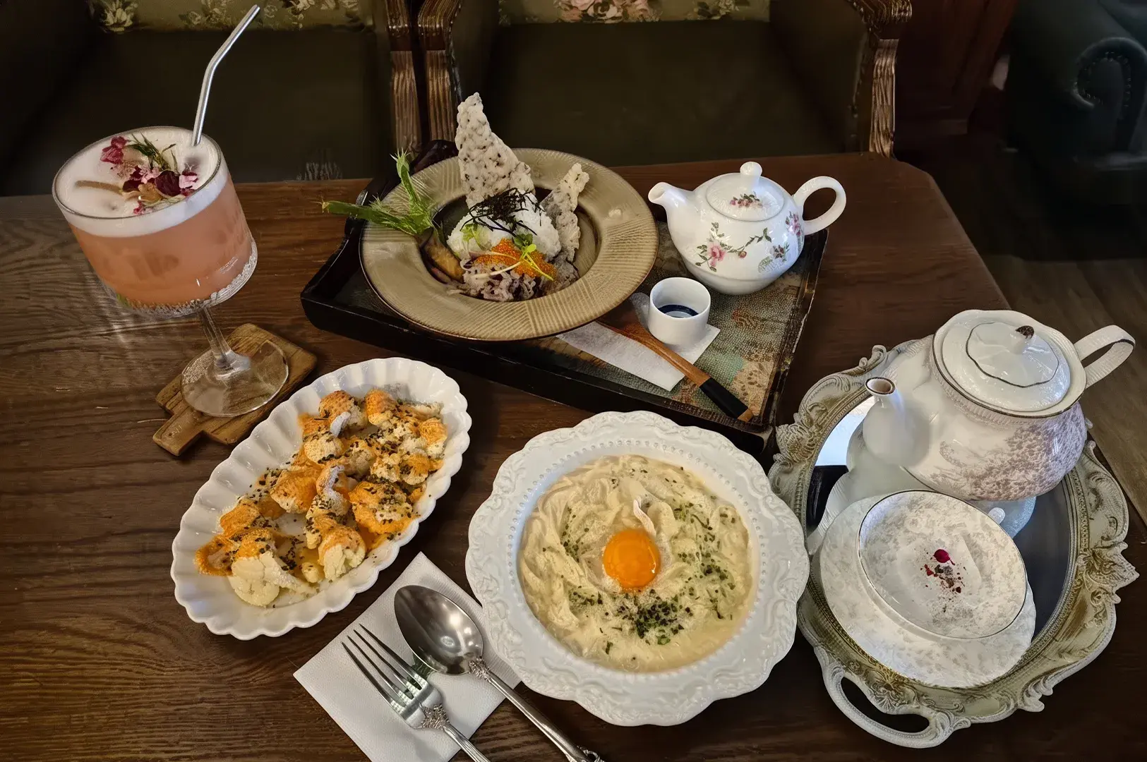 Fusion cafe meal spread with cream pasta, sushi rolls, plated rice, floral tea, and a cocktail.