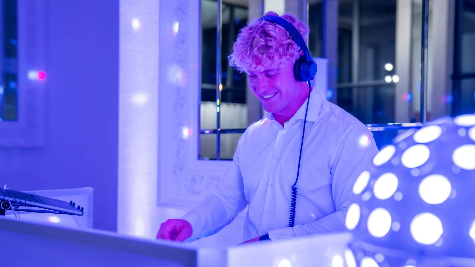 House DJ Nathan White mit Kopfhörern und weißem Hemd legt bei einer Hochzeitsfeier in einem modern beleuchteten Innenraum auf, umgeben von blau-violetter Lichtstimmung und Lichteffekten.
