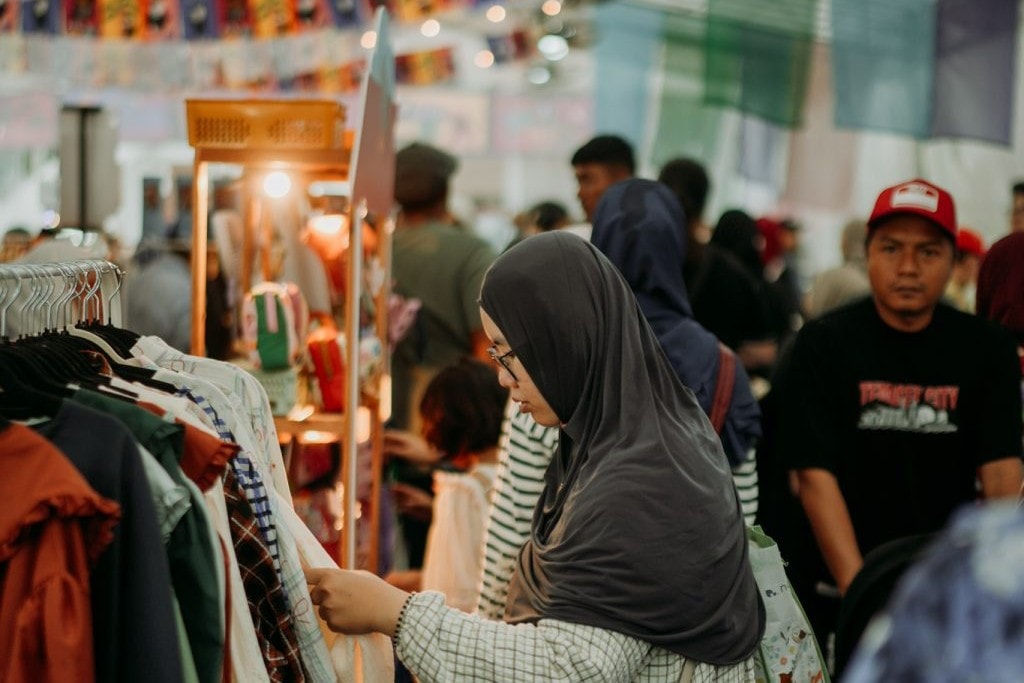 This image shows a woman in a grey hijab browsing through a rack of clothing at a busy indoor market. The background is filled with other shoppers and colorful hanging banners, creating a lively community atmosphere.