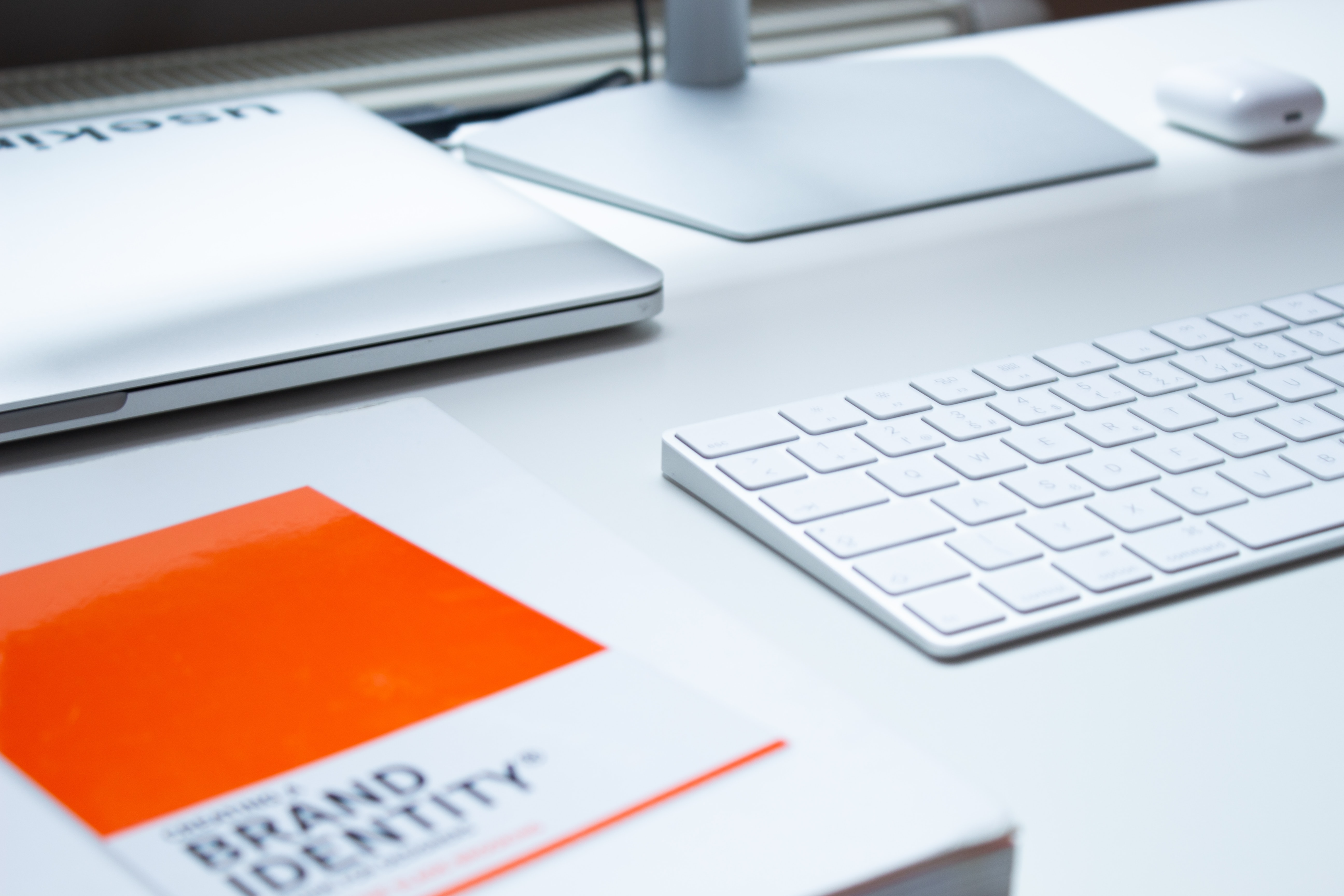Sur une table de couleur blanche sont posés un clavier blanc, un ordinateur portable blanc fermé, un écran d'ordinateur également de couleur blanche ainsi qu'un bloc-notes de couleur orange où il est écrit "Brand identity". L'identité de marque peut faire ou défaire une image de marque.