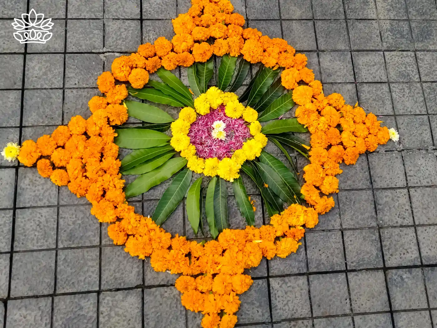 A vibrant outdoor rangoli made with bright orange marigolds and green leaves arranged in a symmetrical pattern on a grey tiled floor, centered with a burst of yellow and pink flowers. Fabulous Flowers and Gifts - Diwali Flowers. Delivered with Heart