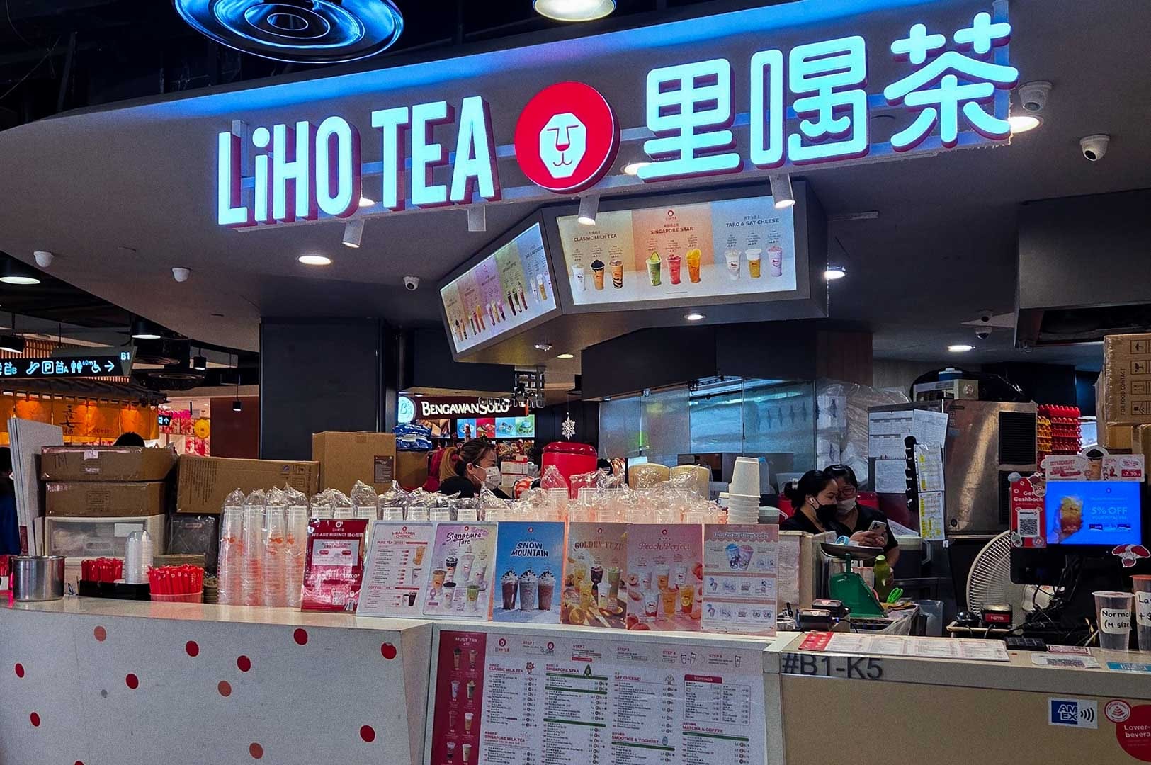 LiHO TEA storefront at Tampines mall with illuminated red lion logo and signage, staff preparing drinks behind a counter lined with clear cups and promotional posters.
