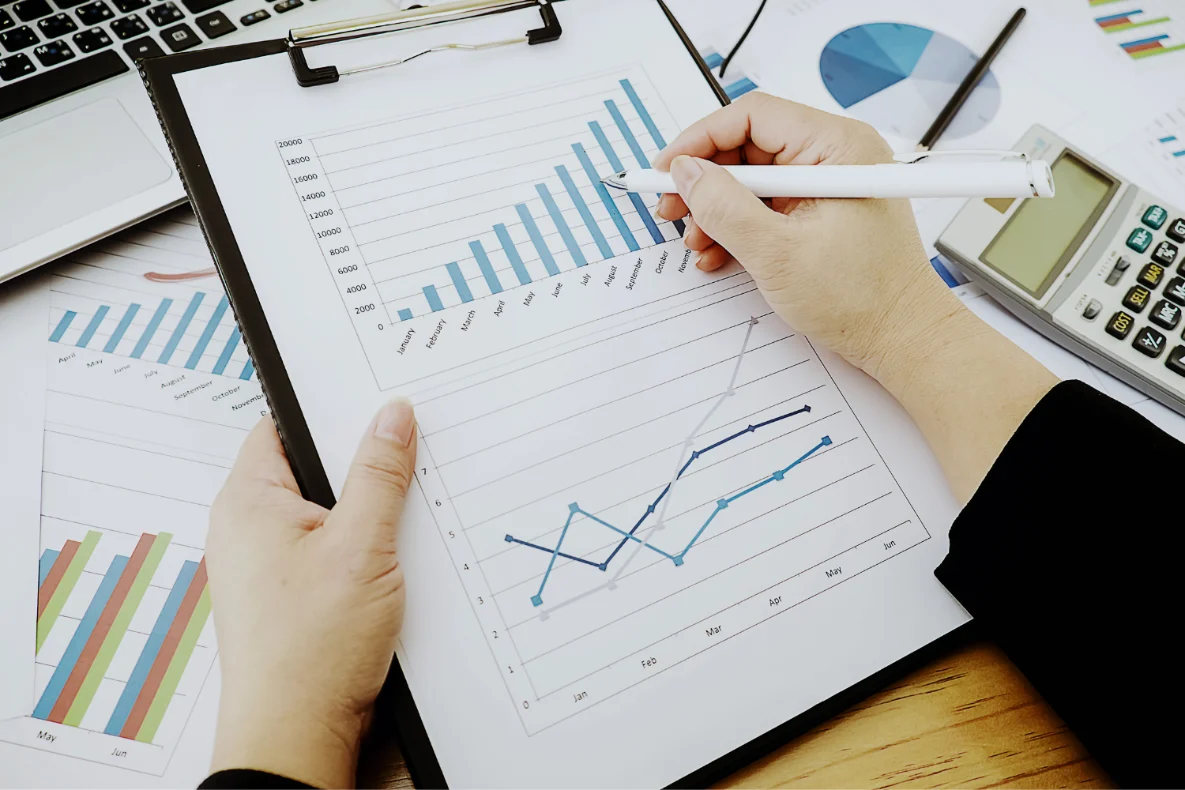 A hand holding a pen points to a bar graph on a clipboard, with other financial charts, a calculator, and a laptop visible on a wooden desk.