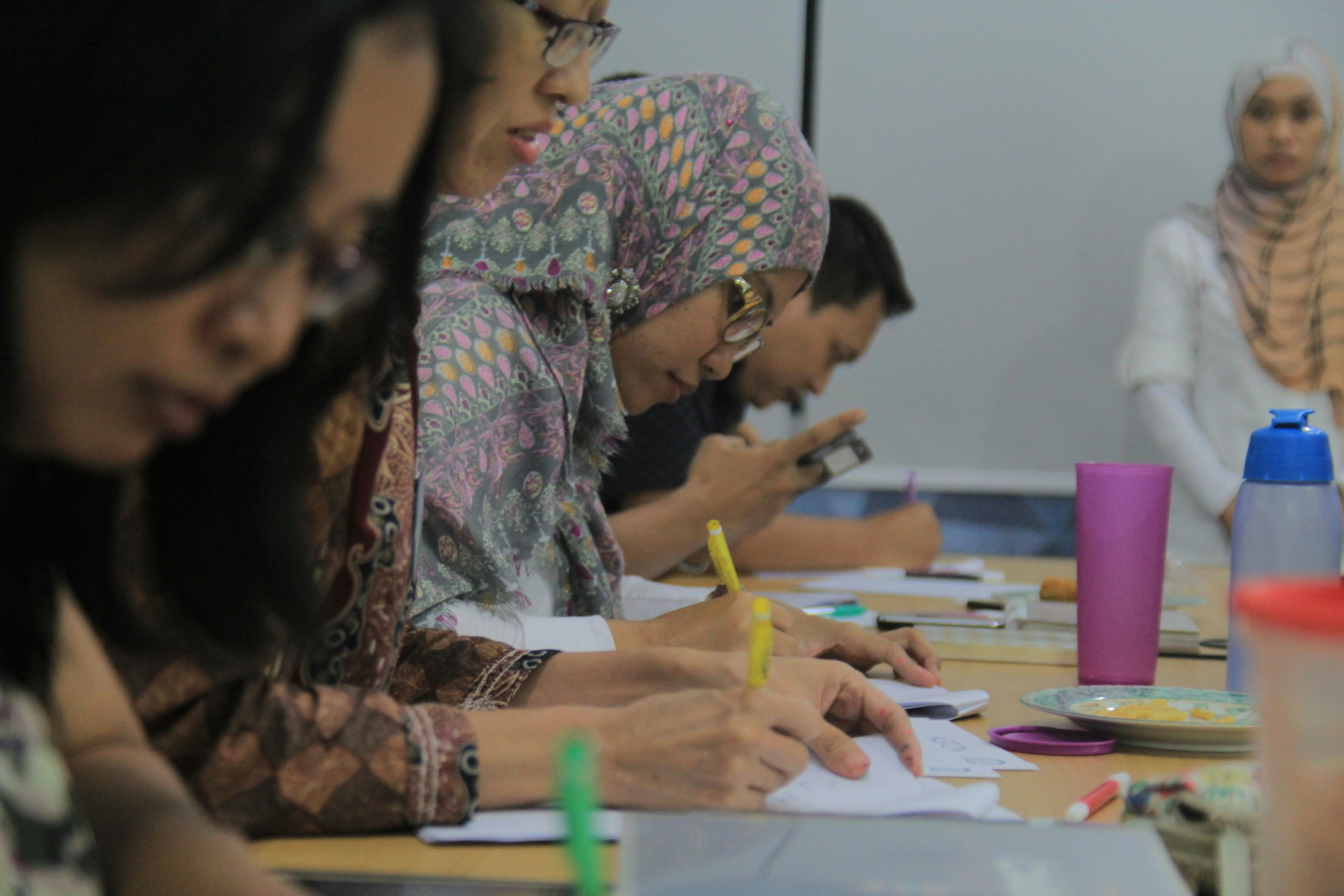 This side-view close-up focuses on a woman wearing a patterned headscarf and glasses who is intently writing notes with a yellow marker during a gathering. The scene suggests a workshop or classroom environment, with other participants visible in the blurred background alongside water bottles and stationery scattered on the table.