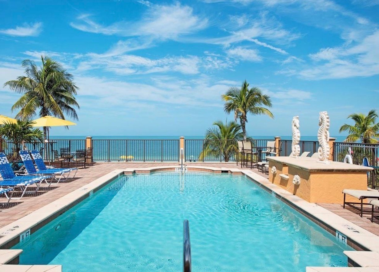 The image depicts a luxurious beach club facility featuring expansive outdoor living spaces with an outdoor dining area that overlooks the stunning Gulf of Mexico. This renovated private beach club offers a perfect setting for both casual family dinners and elegant alfresco dining, surrounded by pristine landscaping and a sparkling swimming pool.