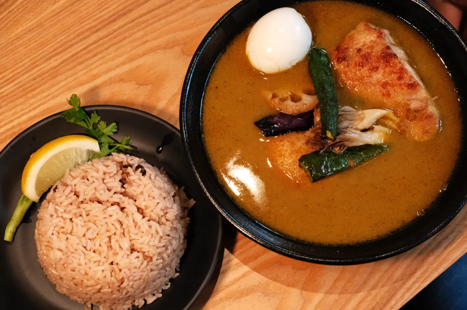 A black bowl of golden curry with egg, browned chicken, eggplant and vegetables sits beside molded rice garnished with lemon and herbs on a wooden table.