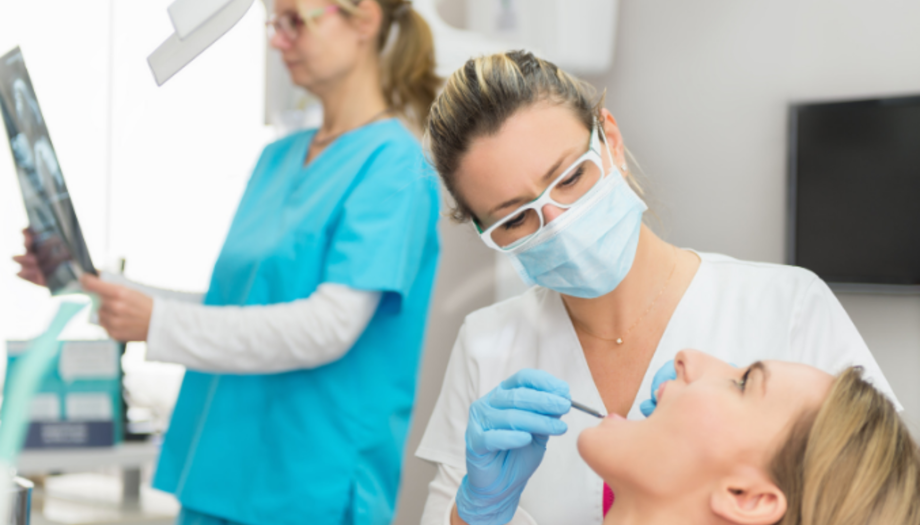 Død Tann? Slik Vet Du Om Tannen Har Sjekket Ut (Men Du Fortsatt Må Inn) 14 Tannlege undersøker kvinnelig pasients tenner mens en tannpleier i bakgrunnen studerer et røntgenbilde.