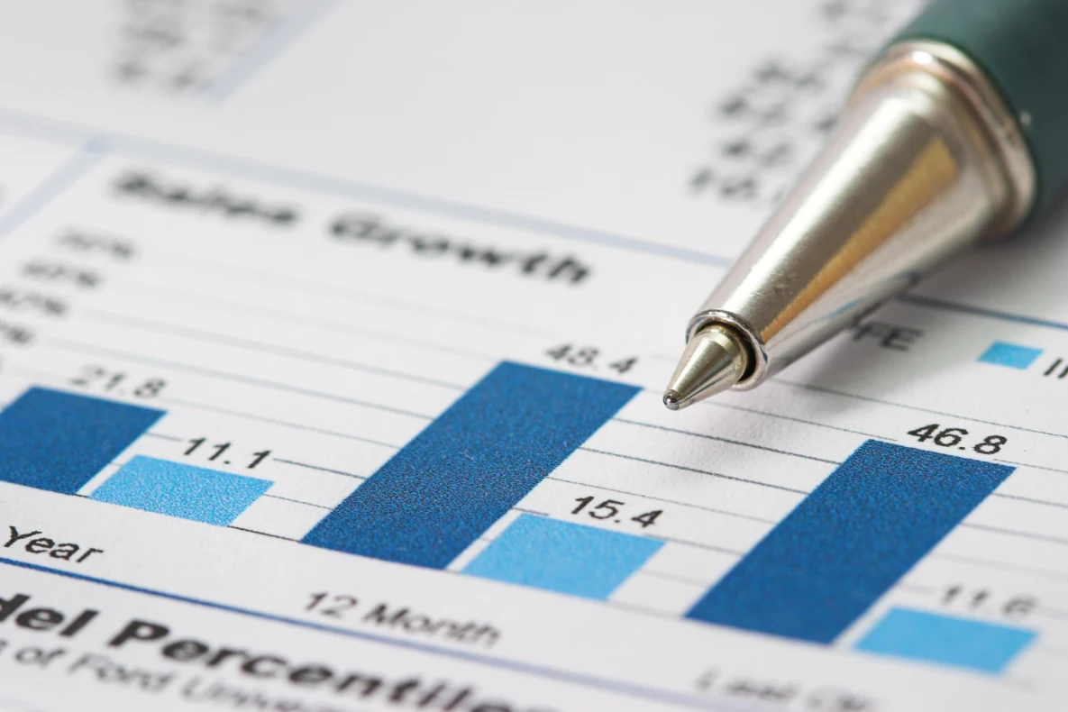 A silver pen resting on a financial report with a bar graph showing sales growth. The graph displays various numerical values and labels like "Year" and "12 Month."