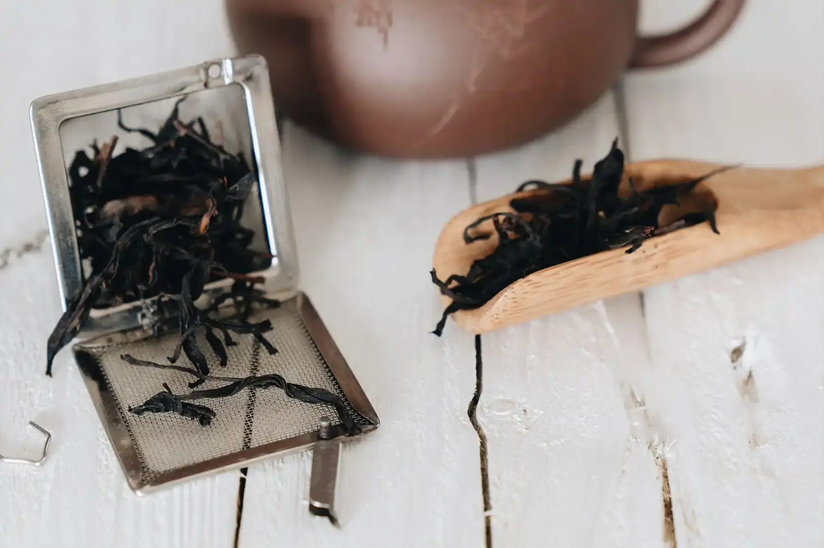 A rustic, close-up shot on a white wooden surface featuring a metal tea infuser filled with dark, dried Lapsang Souchong tea leaves, with a wooden scoop nearby containing more leaves and a blurred clay teapot in the background.