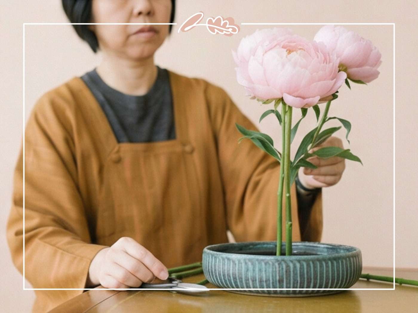 Woman in ochre jacket seated at a wooden table arranging pink peonies in a ribbed teal ceramic ikebana bowl with scissors nearby | Ikebana Practice | Fabulous Flowers & Gifts