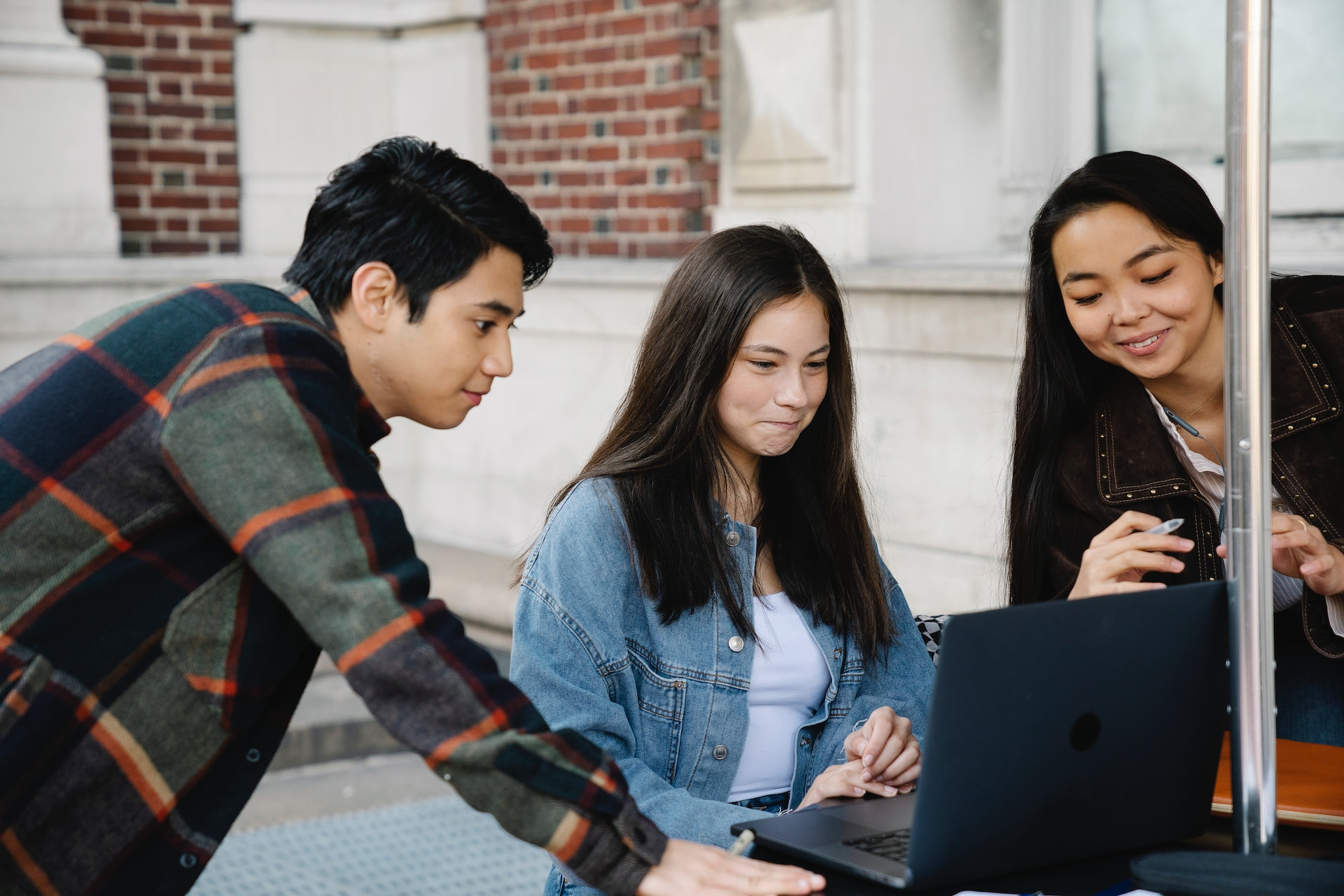 two students lean over the shoulder of a third student in between them who is looking at their laptop screen