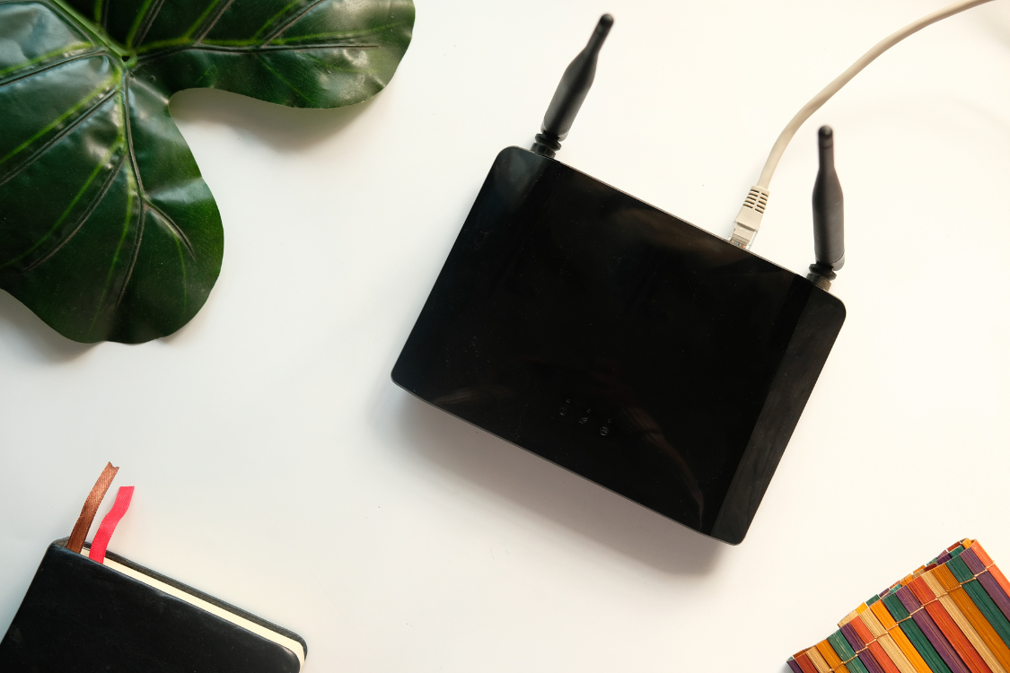 Black Wi-Fi router with connected cable on white desk next to plant and notebook