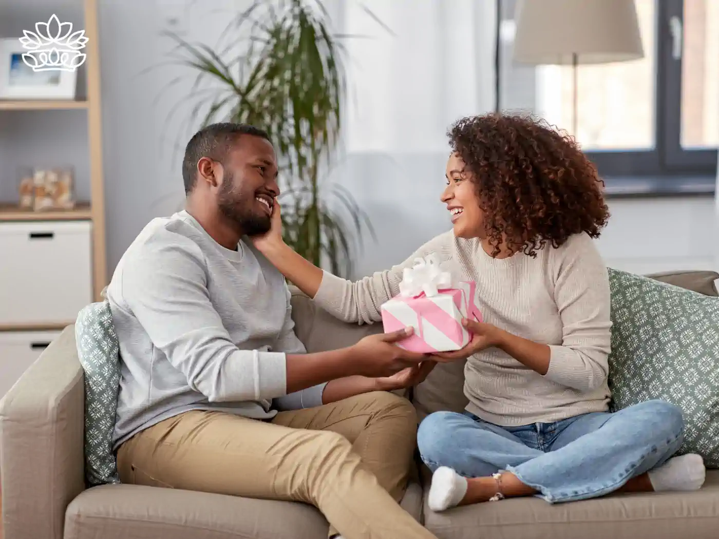 Joyful woman presenting a pink Valentine's Day gift box to a smiling man on a sofa in a modern living room. Fabulous Flowers and Gifts