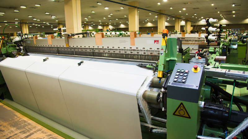 A textile mill with rows of weaving machines processing white fabric under bright overhead lighting.