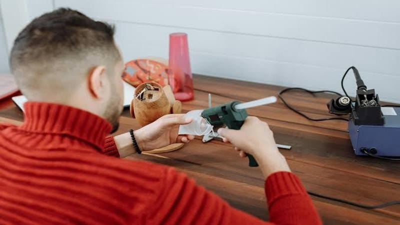 Man in red sweater using a hot glue gun for a wood-based project on a workbench.