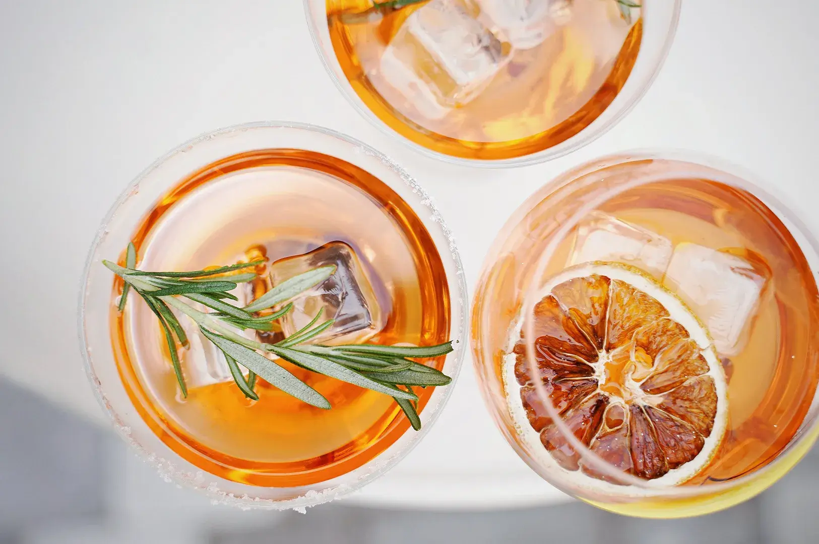Top-down view of three glasses of amber-colored cocktails with ice, garnished with a sprig of fresh rosemary and a dried citrus slice.
