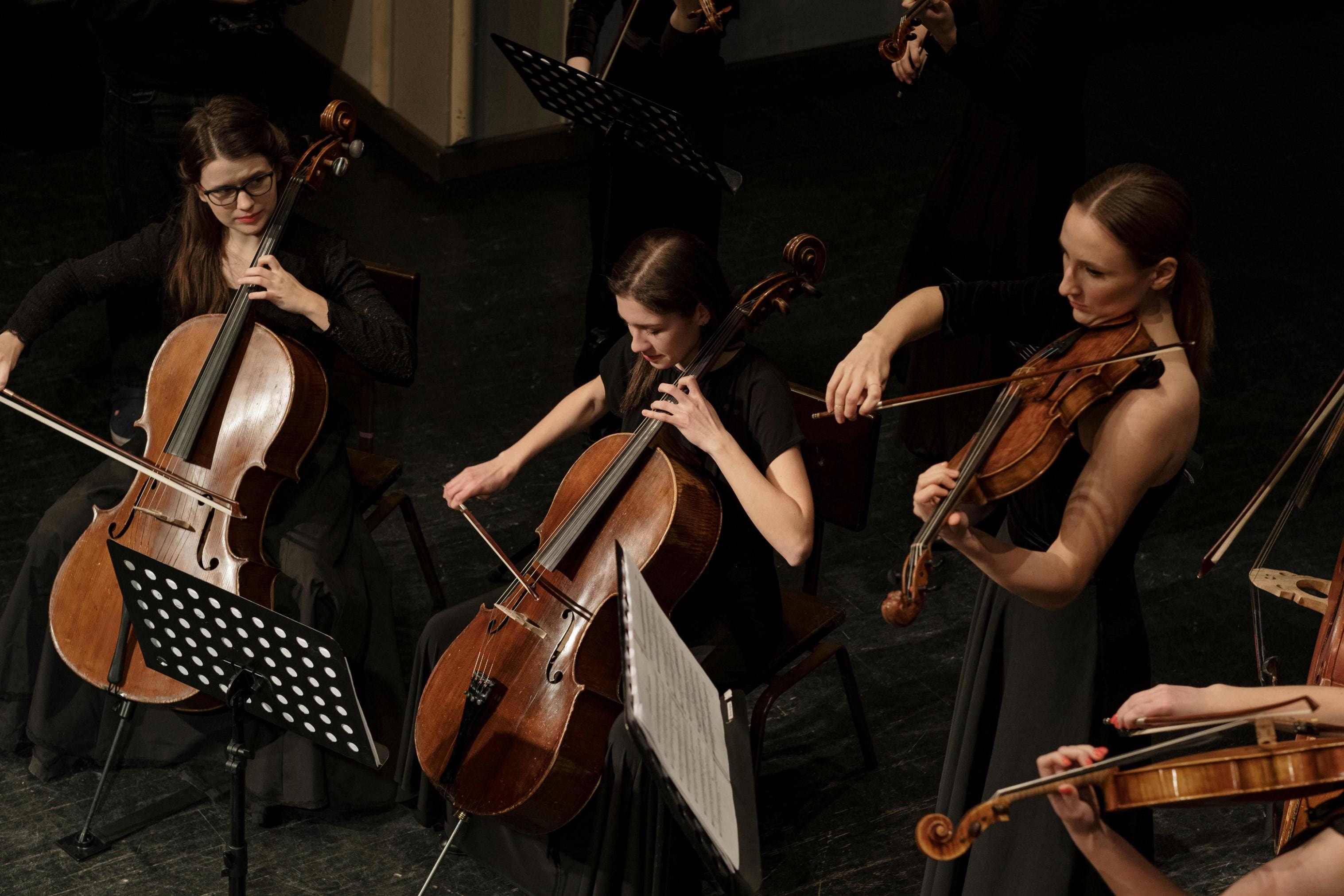 Dressed in formal black attire, a group of female musicians plays string instruments, including cellos and violins, on a wooden stage. The high-angle shot captures their focused expressions as they perform together, with music stands positioned in front of them.