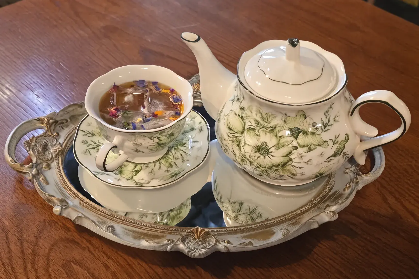Porcelain teapot and iced herbal teacup garnished with edible flower petals on a mirrored tray.