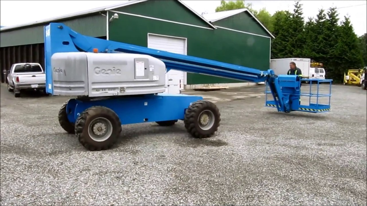 Worker operating a blue Genie telescopic boom lift at an outdoor industrial site, demonstrating the equipment's extended reach and mobility on a gravel surface