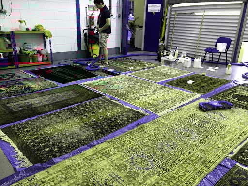 A rug cleaning facility with multiple Oriental and Persian-style rugs laid out on blue tarps for drying or further cleaning. A worker is seen using a machine on one of the rugs, surrounded by cleaning supplies and equipment in a well-lit indoor workspace.