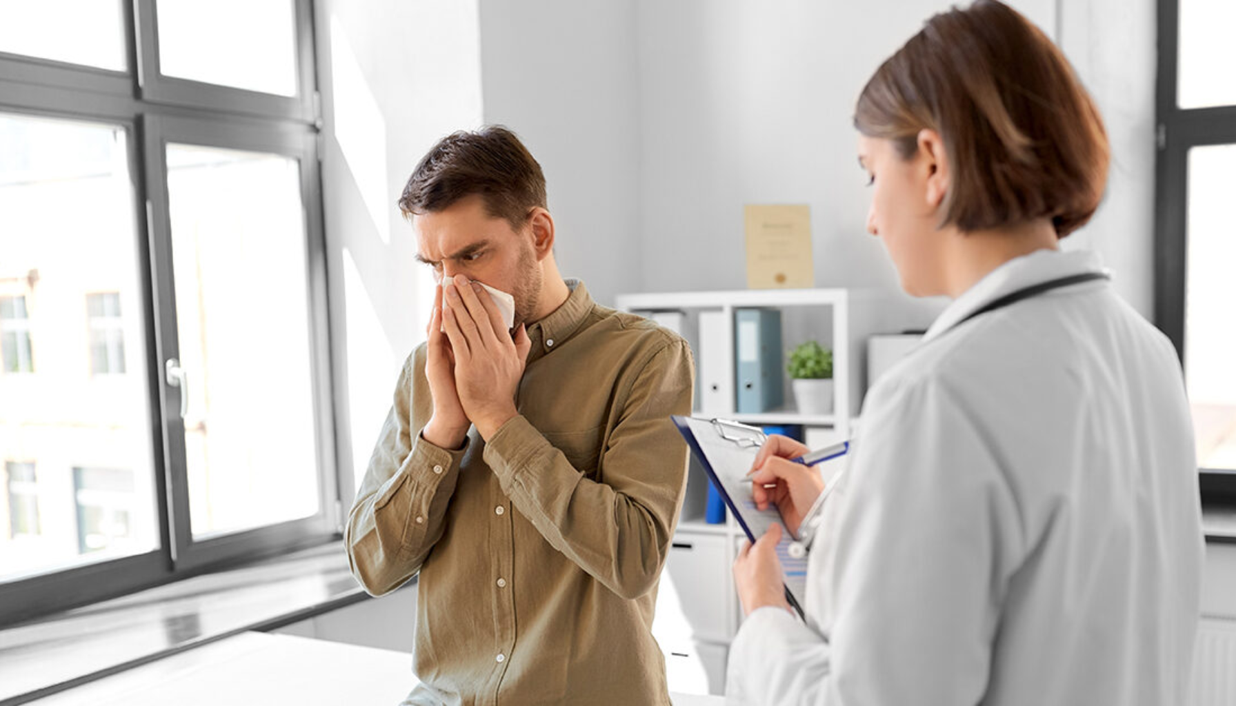 Mann med forkjølelsessymptomer tørker nesen hos lege, mens legen noterer symptomer i en medisinsk journal i et lyst legekontor.