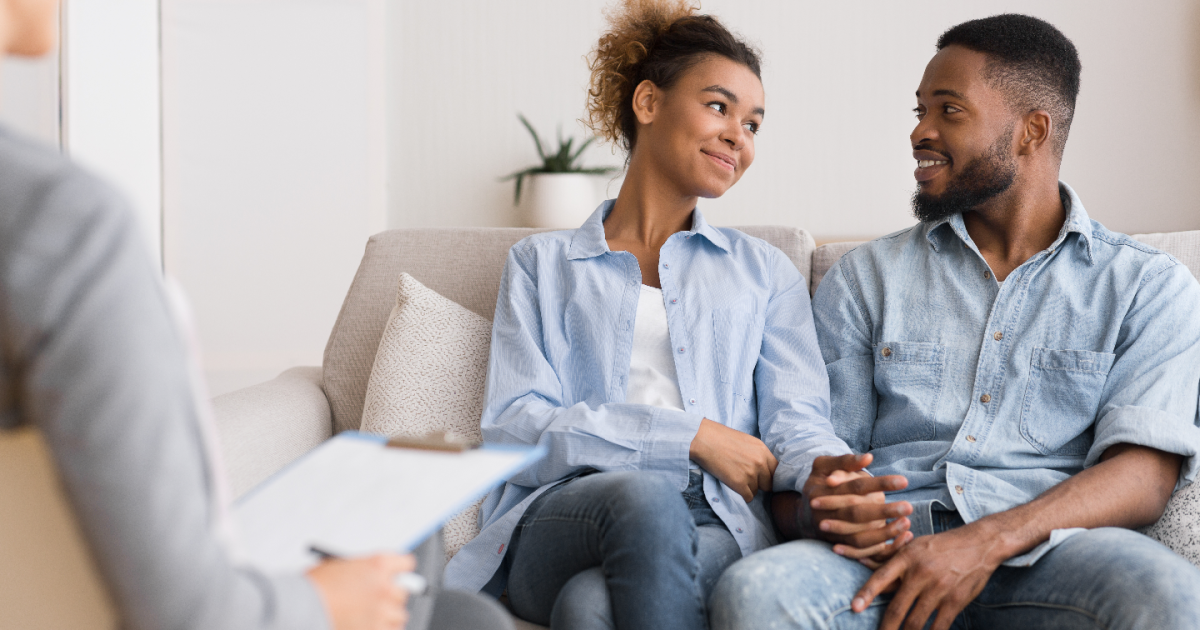 An intimate setting at Loving at Your Best Marriage and Couples Counseling in New York, with a couple sitting close together on a couch, facing a therapist who is attentively listening and taking notes. The room is warmly lit and designed to create a safe and welcoming environment for open communication and relationship building. The couples therapy goals in New York City are clearly met.