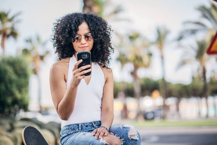 Pretty young woman with dark curly hair and sunglasses reading a text message on her phone. 