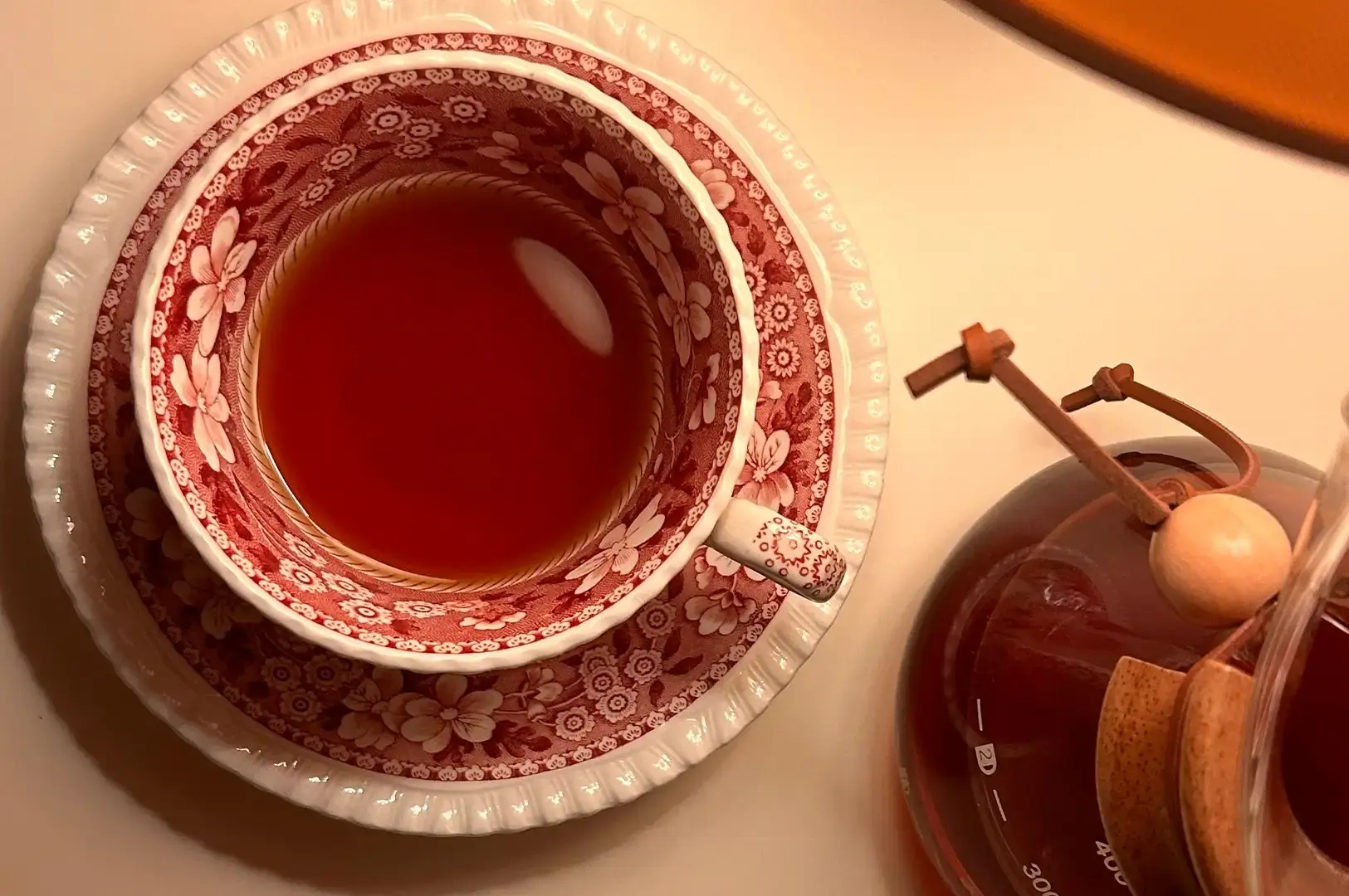 A top-down view shows a delicate vintage teacup adorned with red floral patterns filled with warm amber tea on a matching saucer, placed next to a glass coffee carafe bound with a wooden collar and leather tie.