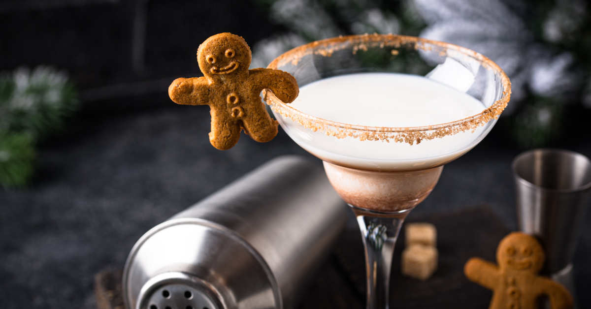 A creamy gingerbread holiday cocktail in a rimmed martini glass garnished with a smiling gingerbread cookie, set beside a stainless-steel cocktail shaker and holiday greenery for a festive Christmas-themed drink presentation.
