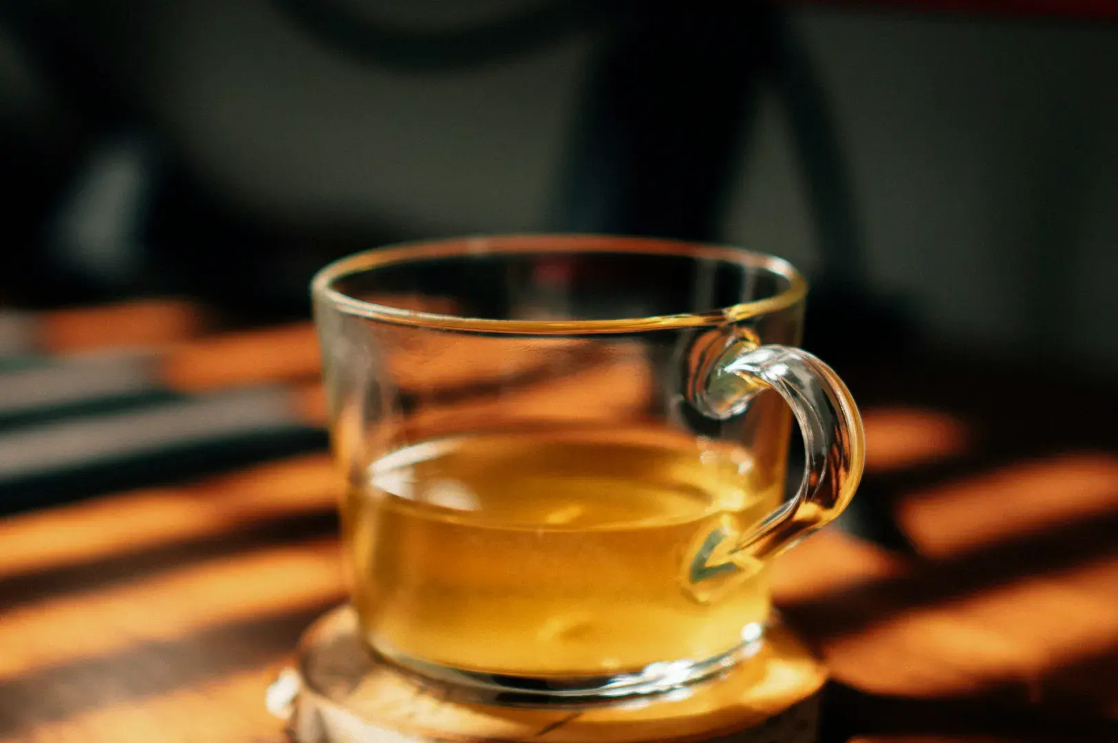 A clear glass cup containing a warm, golden-hued tea is placed on a light wooden coaster. The scene is bathed in warm, directional light, creating distinct, parallel shadows across the wooden table surface beneath the cup, giving the composition a serene and cozy atmosphere.