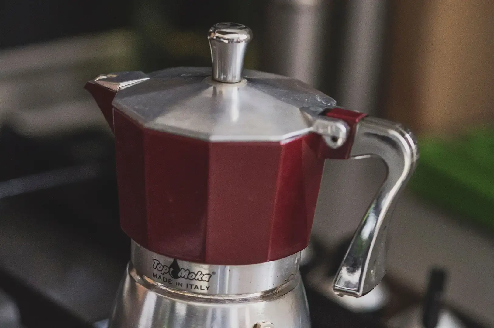 A close-up view of a classic octagonal moka pot coffee maker featuring a deep red upper chamber and a metallic silver base, with the brand name "Top Moka MADE IN ITALY" printed on the silver band.