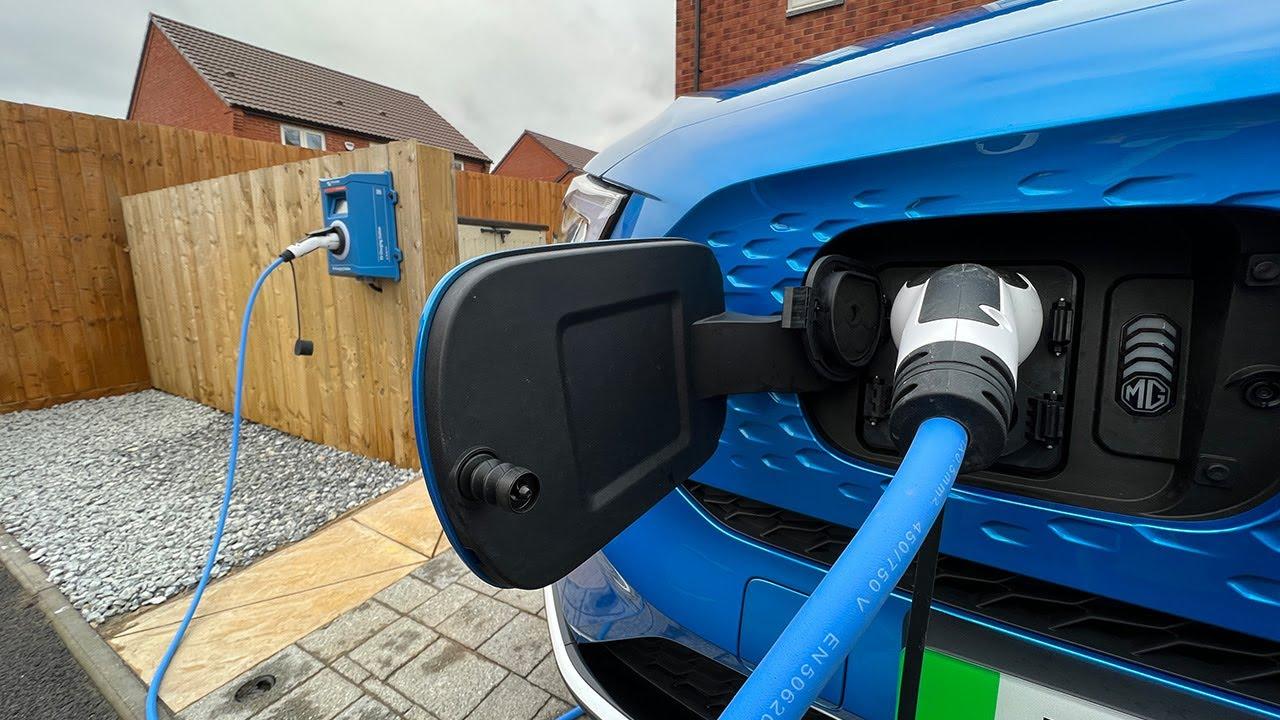 Na zdjęciu widoczny jest dwukierunkowy wallbox zamontowany przy garażu, do którego podłączony jest samochód elektryczny. To nowoczesne rozwiązanie umożliwia ładowanie pojazdów elektrycznych, co wiąże się z niższymi kosztami eksploatacji w porównaniu do tradycyjnych samochodów spalinowych.